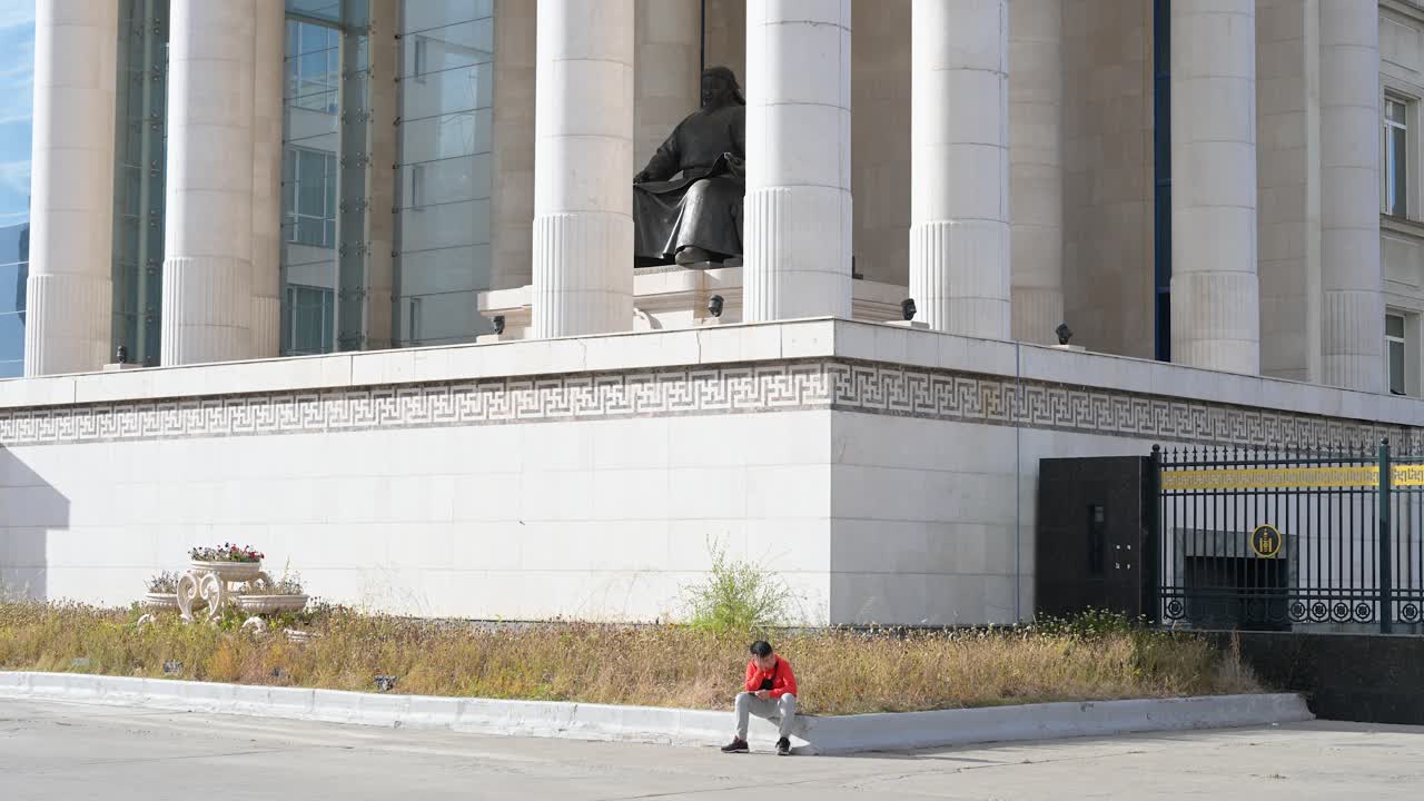 A boy uses a smartphone while sitting in front of the Government Palace in Sukhbaatar Square, Ulaanbaatar, Mongolia, with the Genghis Khan statue positioned at its front center.