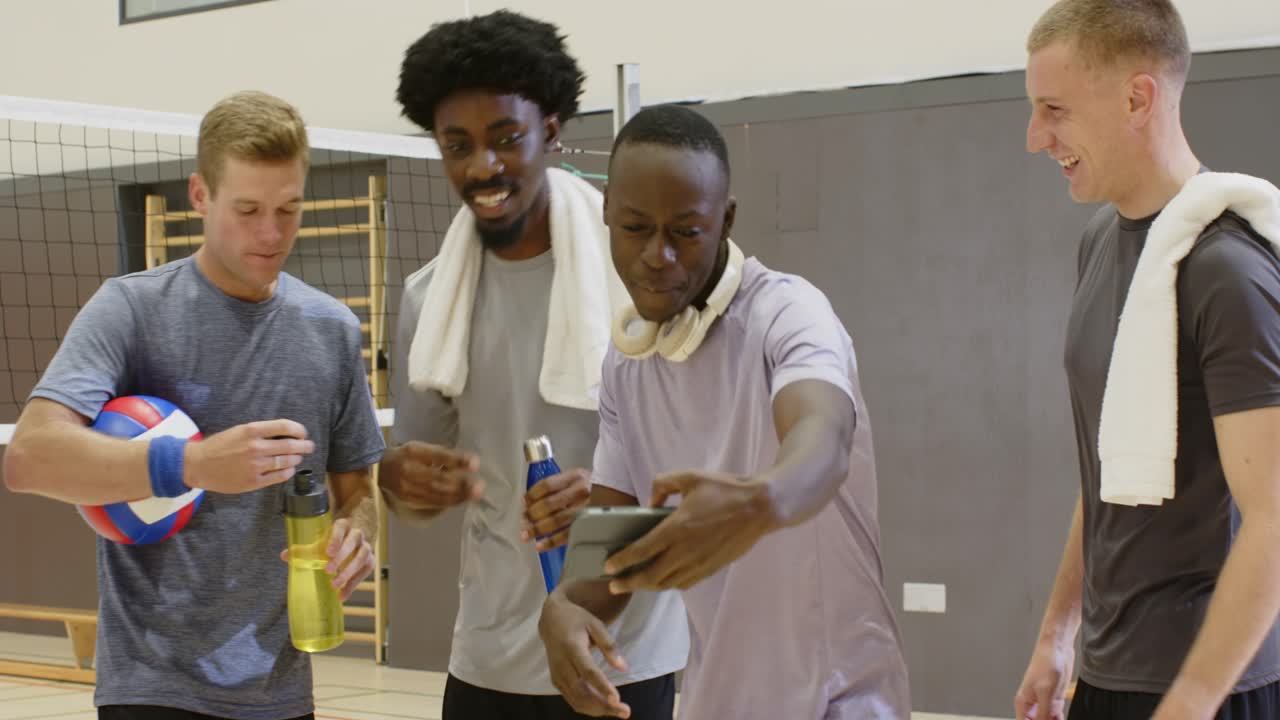 Eine diverse Gruppe von fünf erwachsenen Männern hält einen Volleyball, zeigt ein Smartphone und posiert in der Turnhalle für ein Selfie.