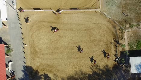 Group-of-horses-and-riders-training-raining-in-outdoor-arena,-aerial-top-down-view