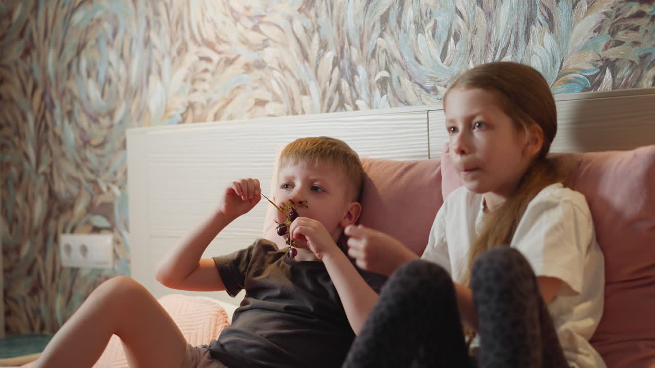 beautiful siblings relaxing on bed enjoying fresh black currant together in cozy room with colorful wall design as they focus on something in background