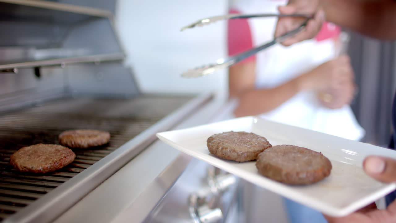 Grilling burgers on barbecue, person using tongs to place patties on plate