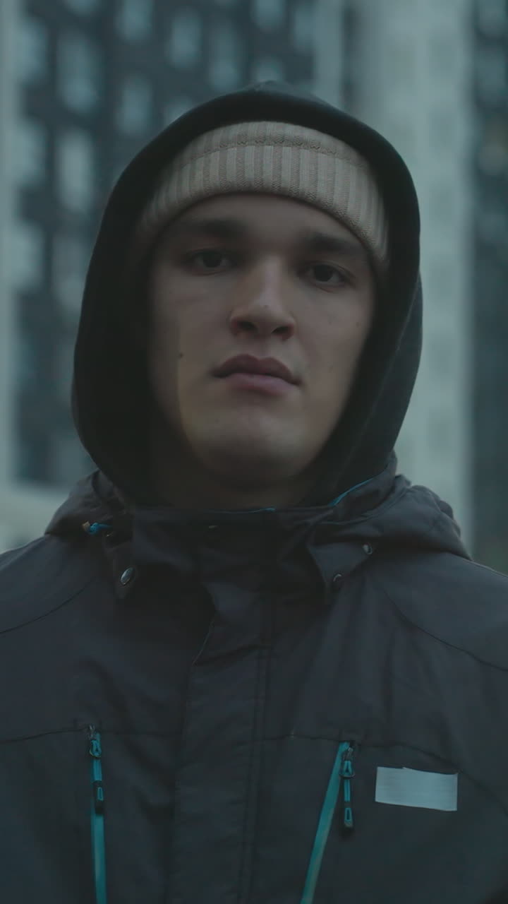 joven aburrido con capucha y gorra haciendo malabarismos con el fútbol durante una sesión de entrenamiento urbano, mostrando una expresión casual contra el telón de fondo de altos edificios residenciales y cielo nublado