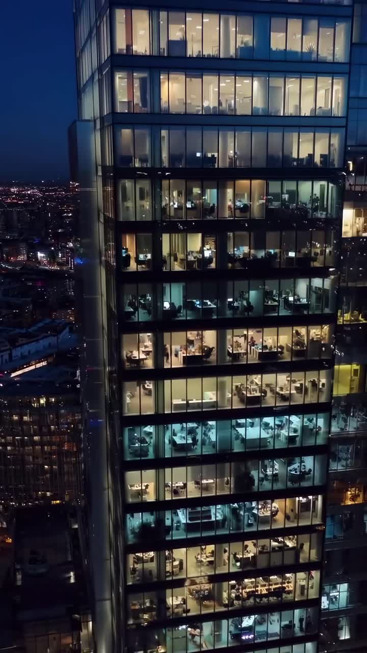 Vídeo aéreo de um arranha-céu iluminado à noite, mostrando a vida de escritório de um ângulo alto