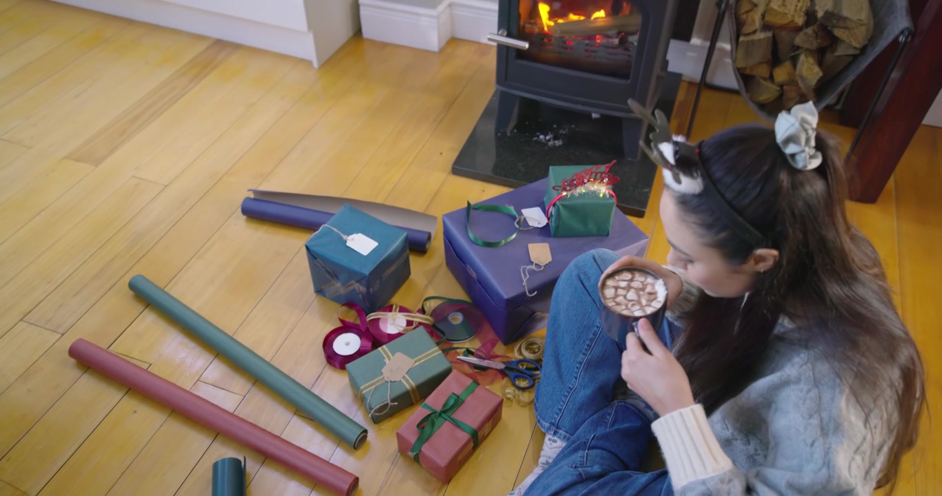 Mujer adulta empieza a organizar papel de regalo, atando una cinta verde, bebiendo de una taza junto a la estufa, con espacio para texto.