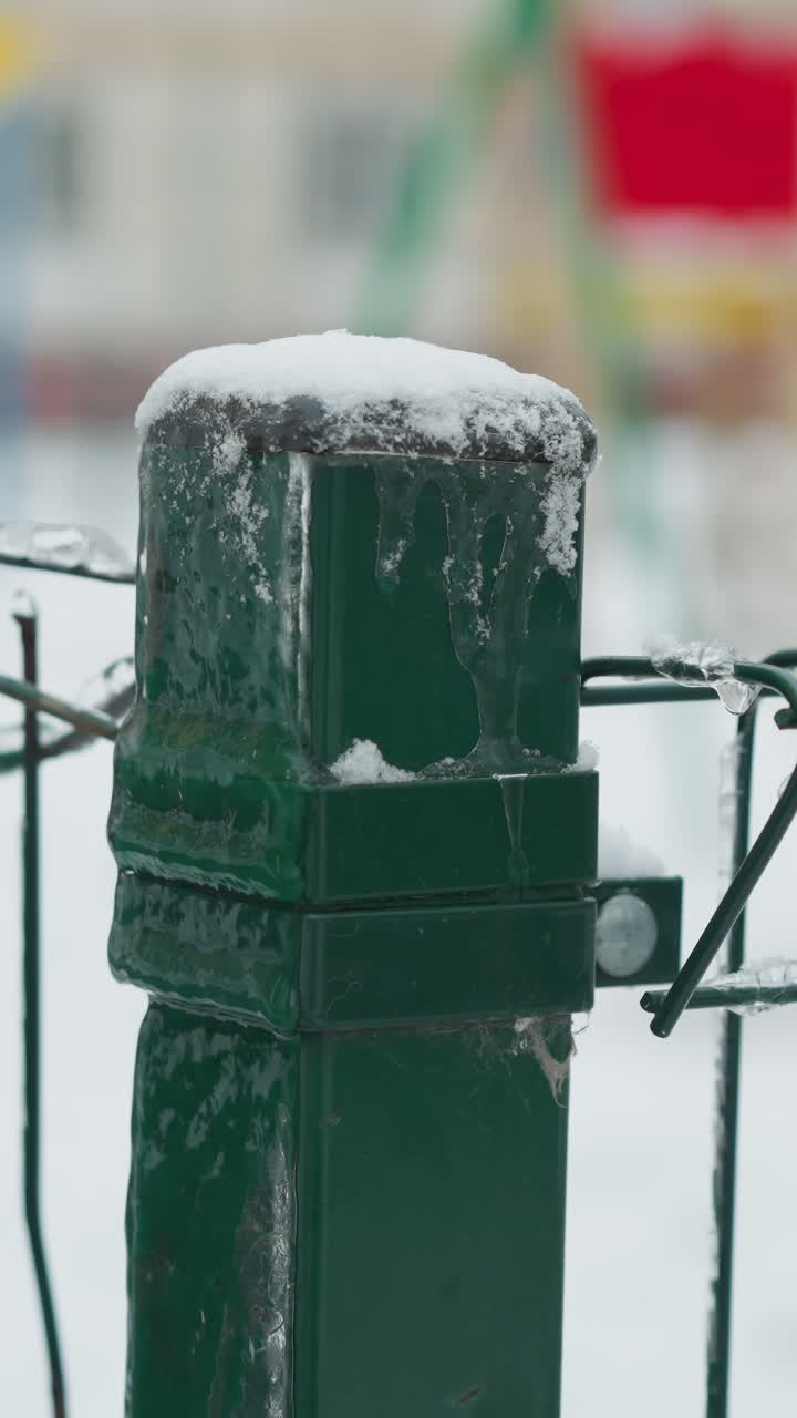 얼음으로 인 초록색 금속 울타리의 클로즈업, 와이어에 붙어있는 아이스클, 흐릿한 눈이 인 놀이터 배경과 멀리 있는 건물