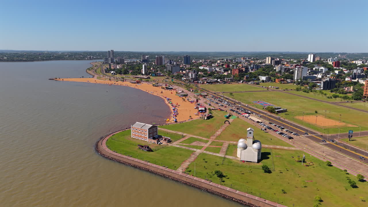 Immerse in a sweeping aerial dolly over Encarnacion’s Playa San Jose in Paraguay, revealing expansive golden sand along a vibrant city skyline under clear, sunny skies