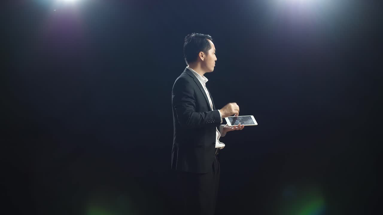 블랙 스크린 스튜디오에서 말하는 동안 태블릿을 들고 가리키는 비즈니스 슈트를 입은 아시아 스피커의 측면 시각