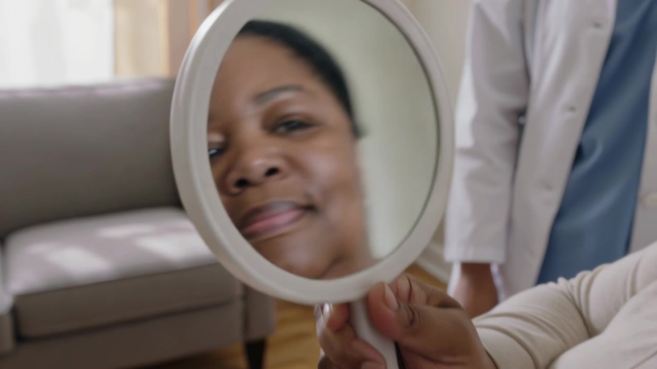 Donna nera sorridente che tiene uno specchio in clinica, controllandosi il viso mentre un medico in camice bianco osserva.