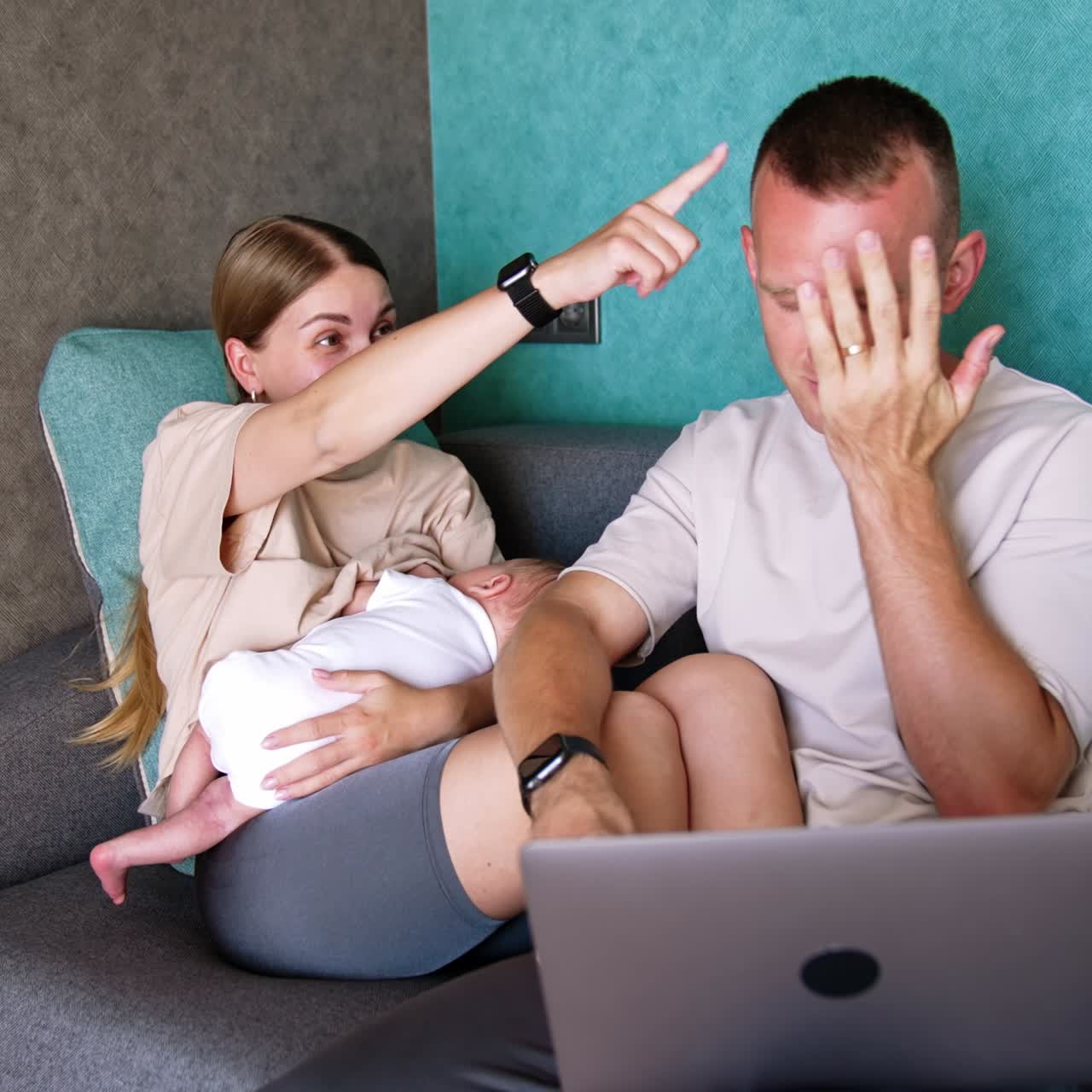 Positive Caucasian couple sit on the sofa. Woman breastfeeding a newborn and man holds a laptop