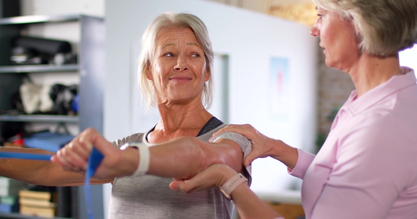 Therapist guiding mature patient in clinic, holding arm while pulling blue band, regaining motion