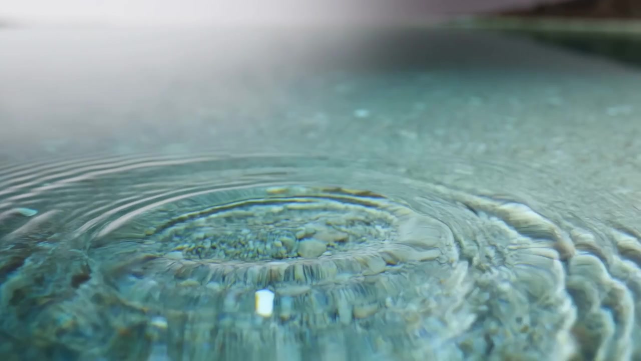 Gotas repetidas perturban una superficie de agua azul verdosa, formando pequeñas salpicaduras y anillos concéntricos que se extienden por un suelo texturizado bajo una luz suave en una composición de primer plano bajo.