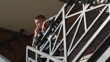 student in black and white shirt smokes cigarette while leaning on metal walkway railing of brick building under bright sunlight with geometric shadows and urban structure in background