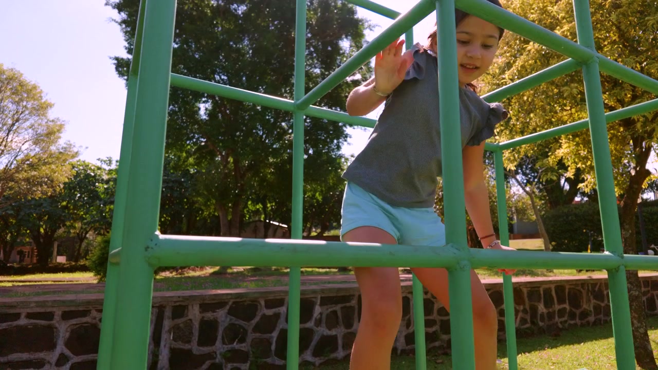 A young girl skillfully climbs a green playground structure in a sunlit park. Ideal for family or outdoor activity themes.