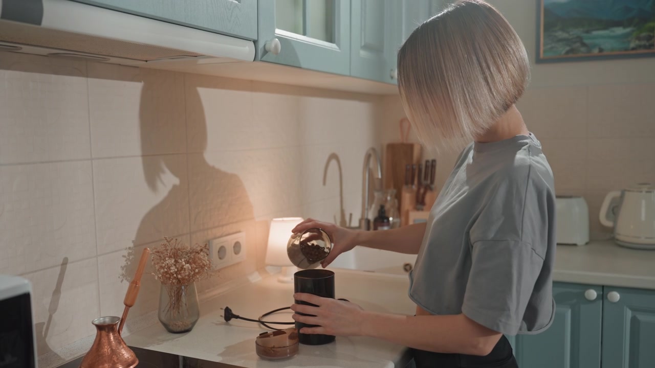 Femme préparant du café sur le comptoir de la cuisine, moulant des grains et versant le marc dans un verre tandis qu'une lampe chaude projette une ombre. Routine matinale intime montrant les mains, une bouilloire en cuivre, un pot en céramique, lente et concentrée.