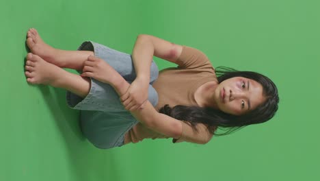 full body of young asian woman victim of violence with bruise on body sits in the green screen background studio and looks into the camera painfully