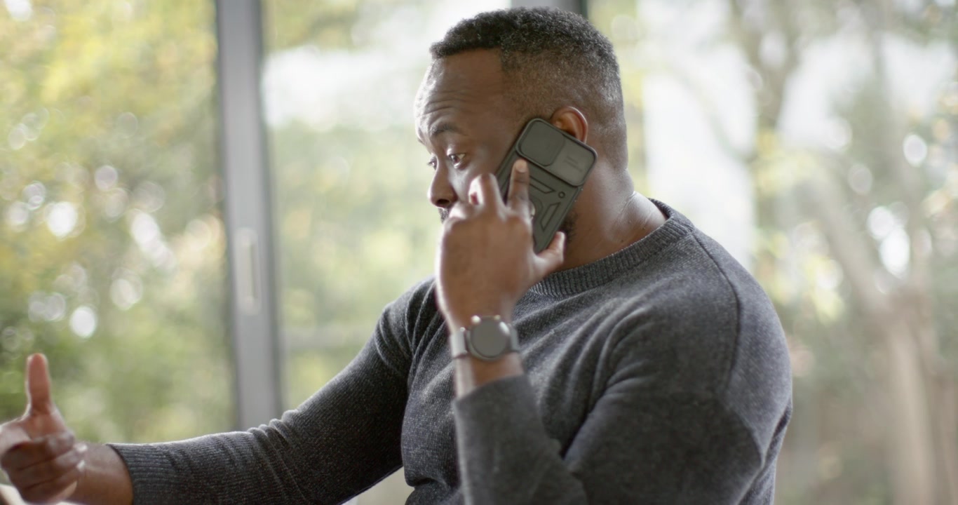 African American man at table answering work call on phone, reaching for tablet, checking watch