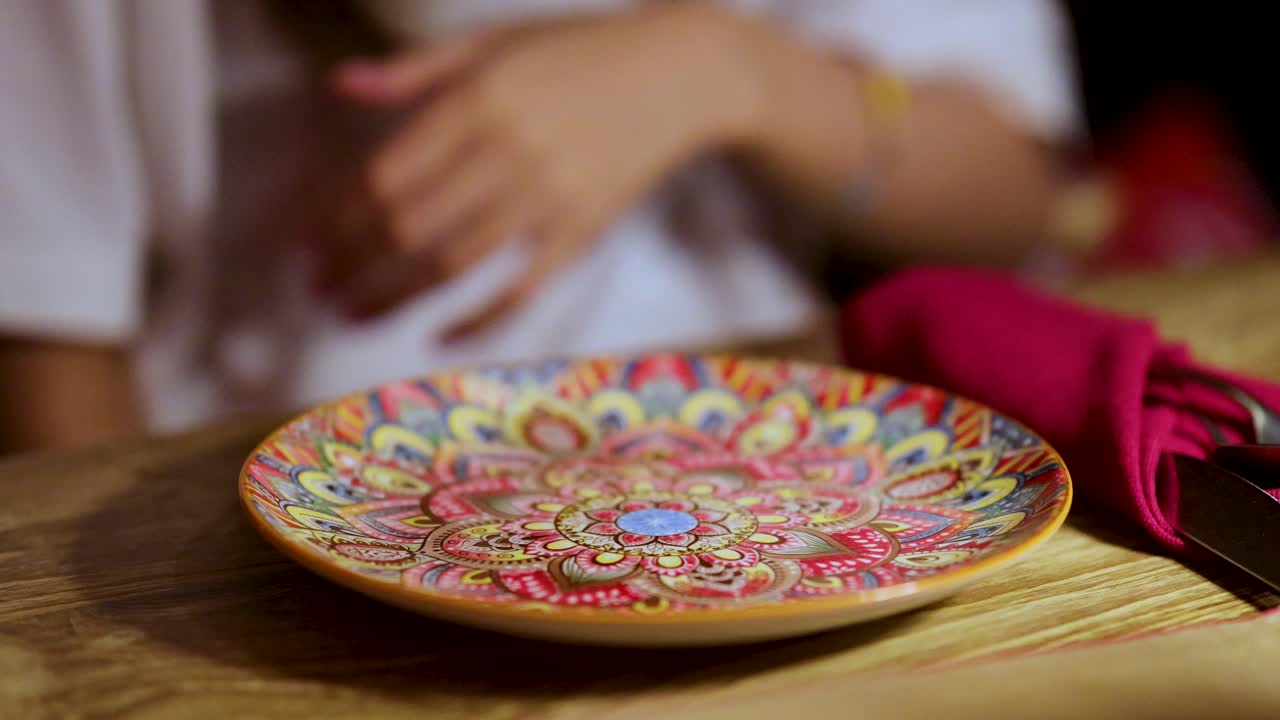 Woman in white shirt waits at wooden table with decorative plate, red napkin, natural lighting