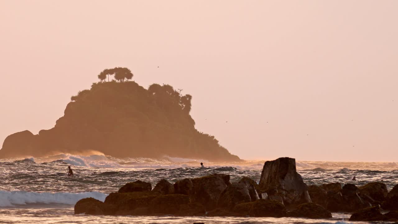 A cinematic scene of a rocky beach bathed in golden sunset light in Weligama, Sri Lanka. A lone surfer walks along the shoreline, silhouetted against the glowing sky and ocean.