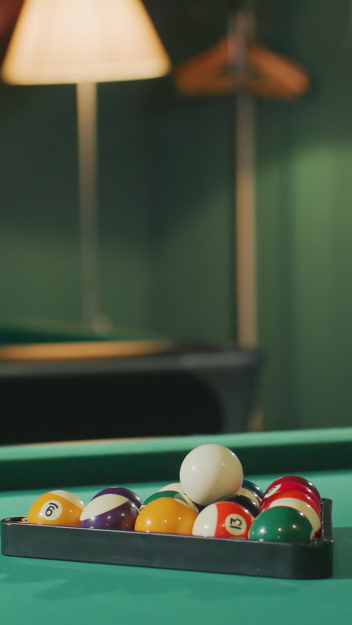 Vibrant billiard balls arranged in triangle rack on green pool table under warm lighting in cozy game room. Classic cue sport setup with focus on white cue ball and numbered balls in dim ambiance
