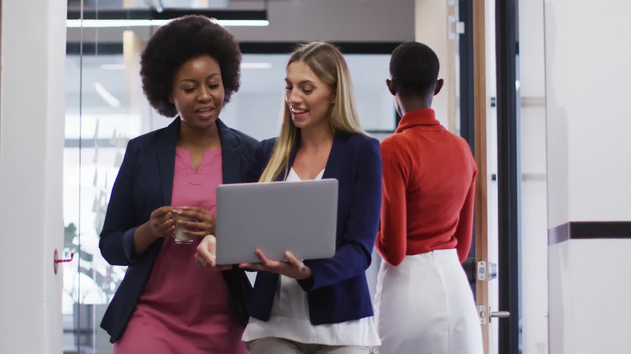 Two diverse female office colleagues with laptop discussing together while walking in office