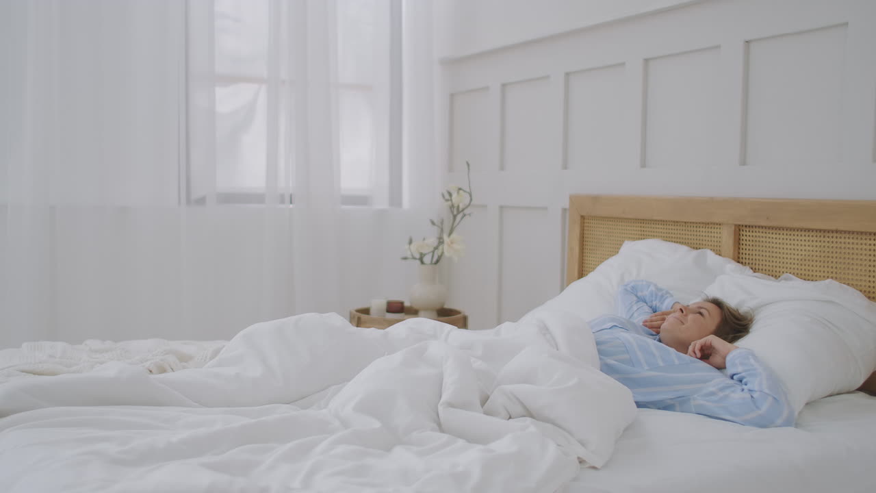 행복한 아름다운 여성은 침실에서 침대에서 미소 짓고 팔을 펴고 깨어납니다. 젊은 여성은 집에서 휴식을 취합니다. 생활 방식 집에서 여성 개념.