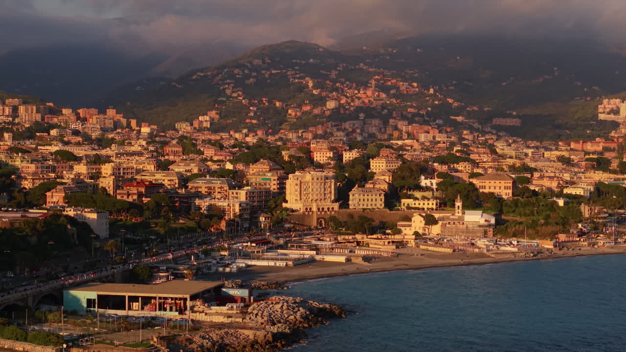 Drone che orbita sopra la costa di Genova mostrando spiaggia, edifici e montagne al tramonto.