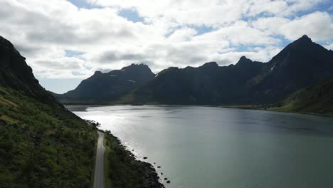 Ascending-aerial-drone-footage-over-scenic-road-in-Lofoten,-Norway-showing-the-ocean-sea-and-the-surrounding-mountains-including-the-power-electricity-cables