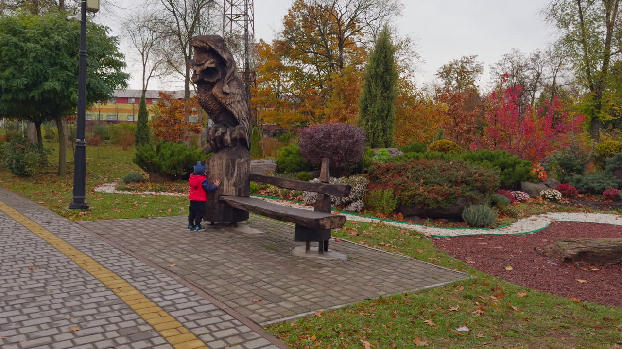 Lovely little toddler standing near the wooden sculpture outdoors. Beautiful autumn park at backdrop.
