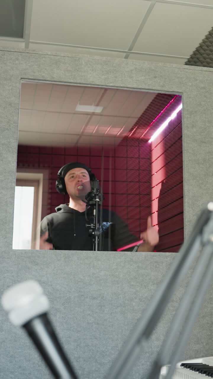 A man dressed in black stands in front of a studio microphone, wearing headphones as he moves his hands while singing in a red soundproofed room. The image also includes a speaker in the background