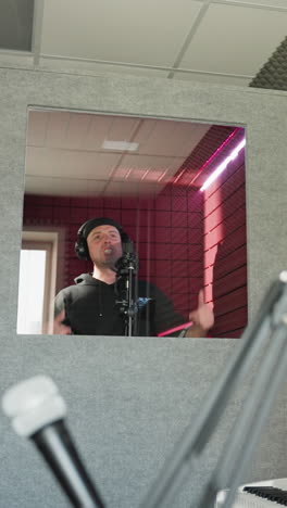 un hombre vestido de negro se encuentra frente a un micrófono de estudio, con auriculares mientras mueve las manos mientras canta en una habitación a prueba de sonido roja. la imagen también incluye un altavoz en el fondo