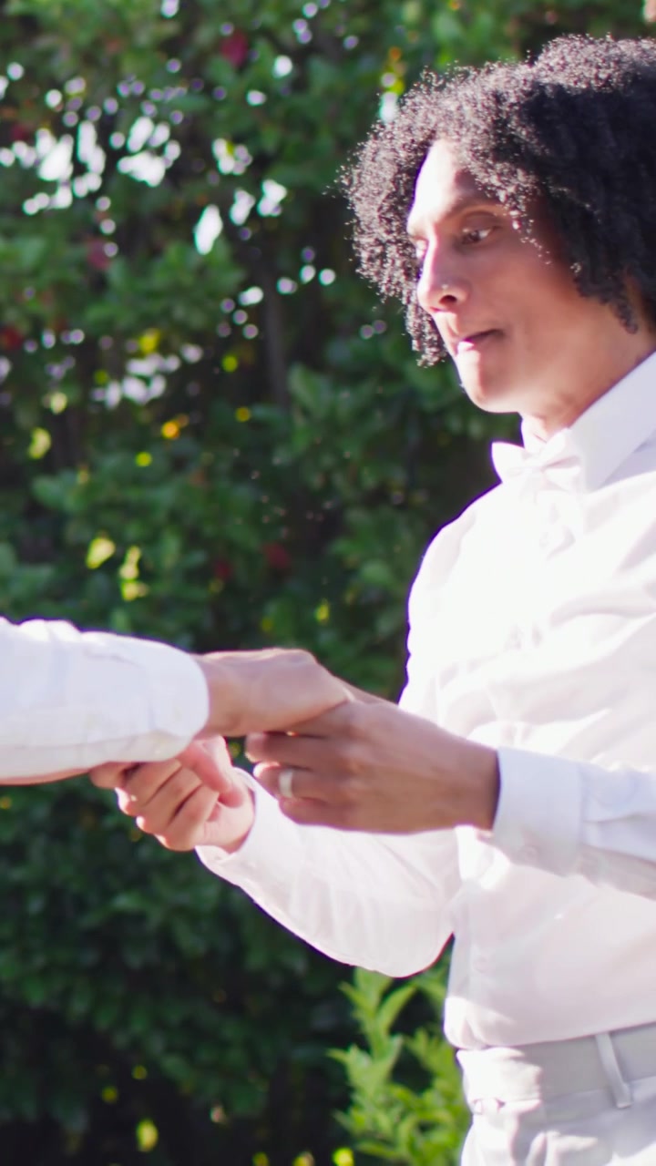 Vertical video: Placing hand on partner's waist, curly man sliding ring in garden, wearing bow tie