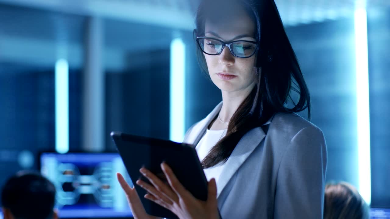 una joven empleada del gobierno usa una tableta en el centro de control del sistema. en el fondo sus compañeros de trabajo están en sus espacios de trabajo con muchas pantallas que muestran datos valiosos.