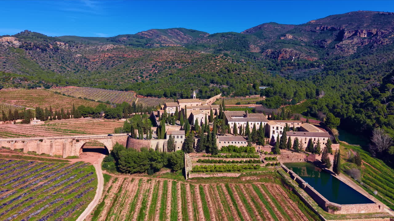 Monastero di Porta Coeli immerso in una valle boscosa a Valencia, Spagna, circondato da vigneti a terrazze, cipressi, un acquedotto in pietra e uno stagno riflettente.