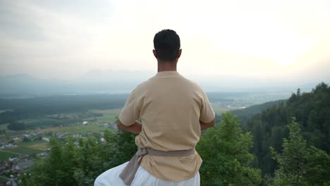 Push-in-shot-of-Indian-man-doing-hatha-yoga-standing-at-the-edge-of-a-stone-castle-wall-in-the-morning-sun-at-sunrise-overlooking-the-valley-bellow-with-fields-and-woods