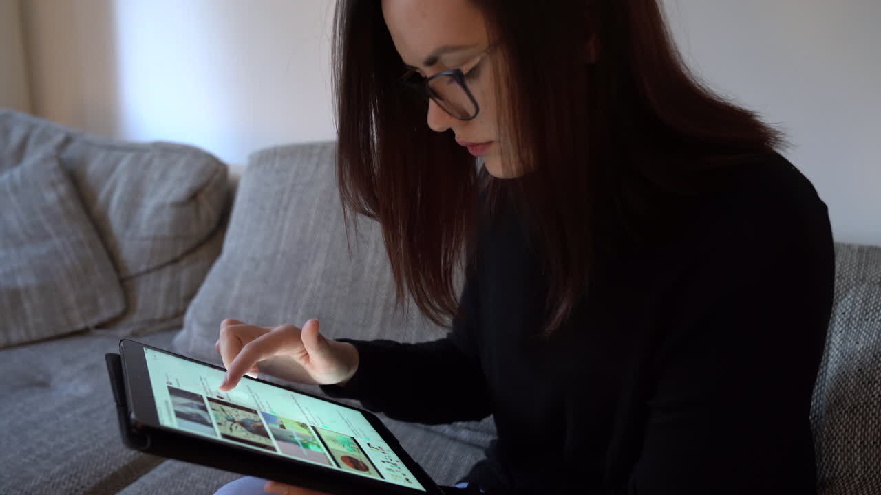 mujer joven seria sentada en el sofá desplazándose en una tableta buscando en internet toma de mano