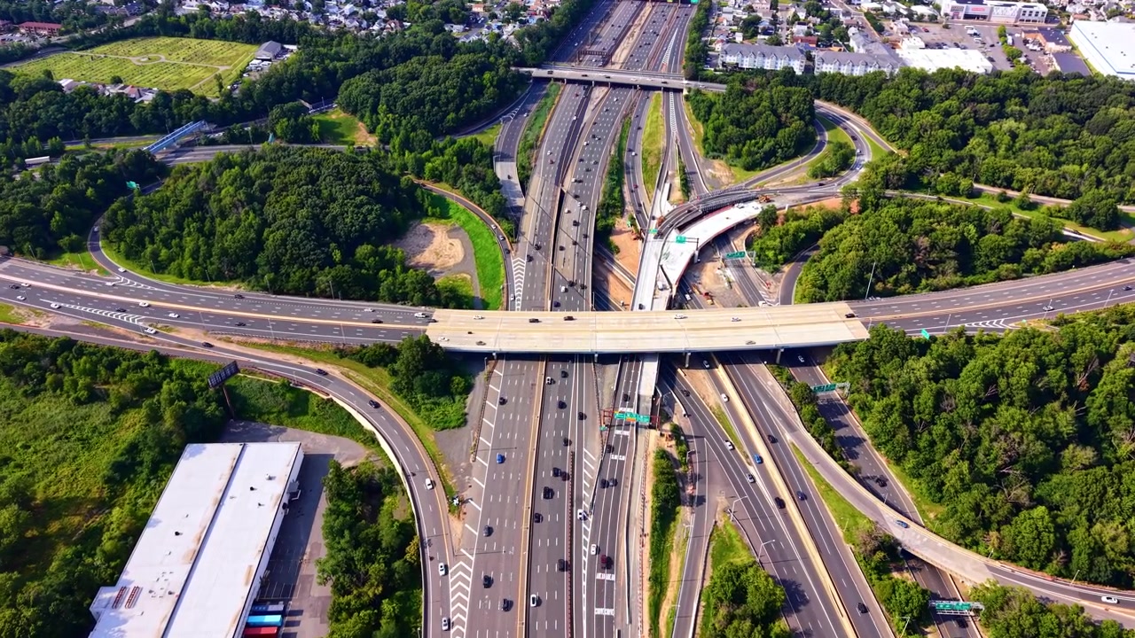 Aerial view of highway interchange near suburban town, New Jersey. Overhead drone shot of multilane roads, overpass bridge, warehouse and green tree cover. Garden state parkway