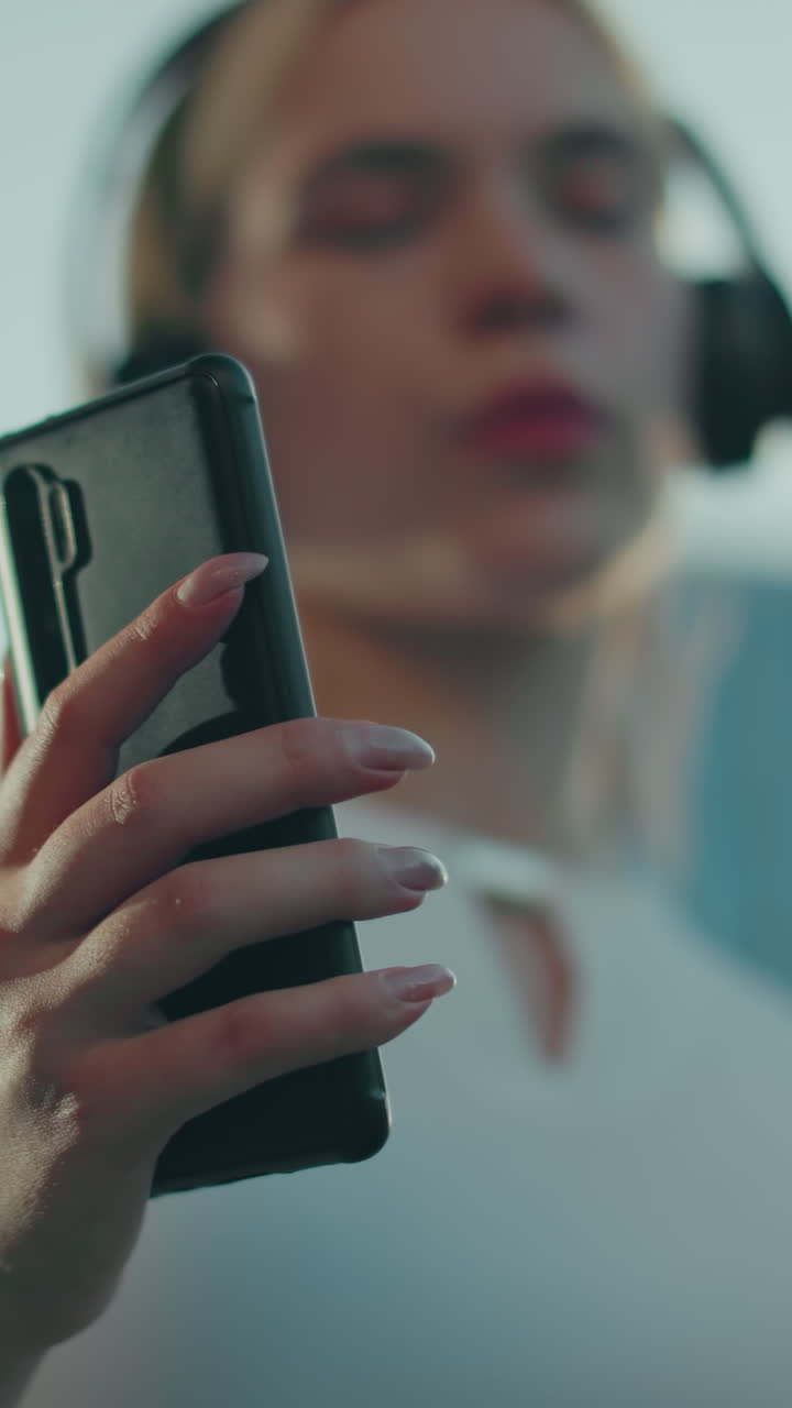 Close up of lady holding phone while wearing headphone and white top, slightly blurred as she dances under bright sky, focus on hand and phone