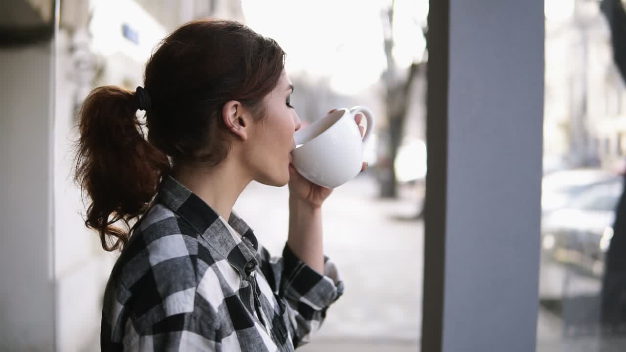 Vista de hermosas chicas de pie cerca de las ventanas. Tomando un té en una taza blanca. Vista lateral. Fondo borroso.