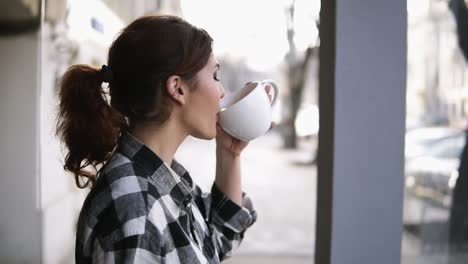 Gorgeous-girls-view-standing-near-the-windows.-Drinking-a-tea-with-a-white-cup.-Side-view.-Blurred-background