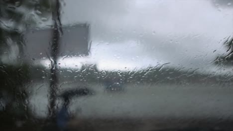 Looking-out-of-a-window-covered-in-water-droplets-to-see-a-person-sitting-under-an-umbrella-in-the-pouring-rain-along-the-Mandovi-River,-Panjim,-India