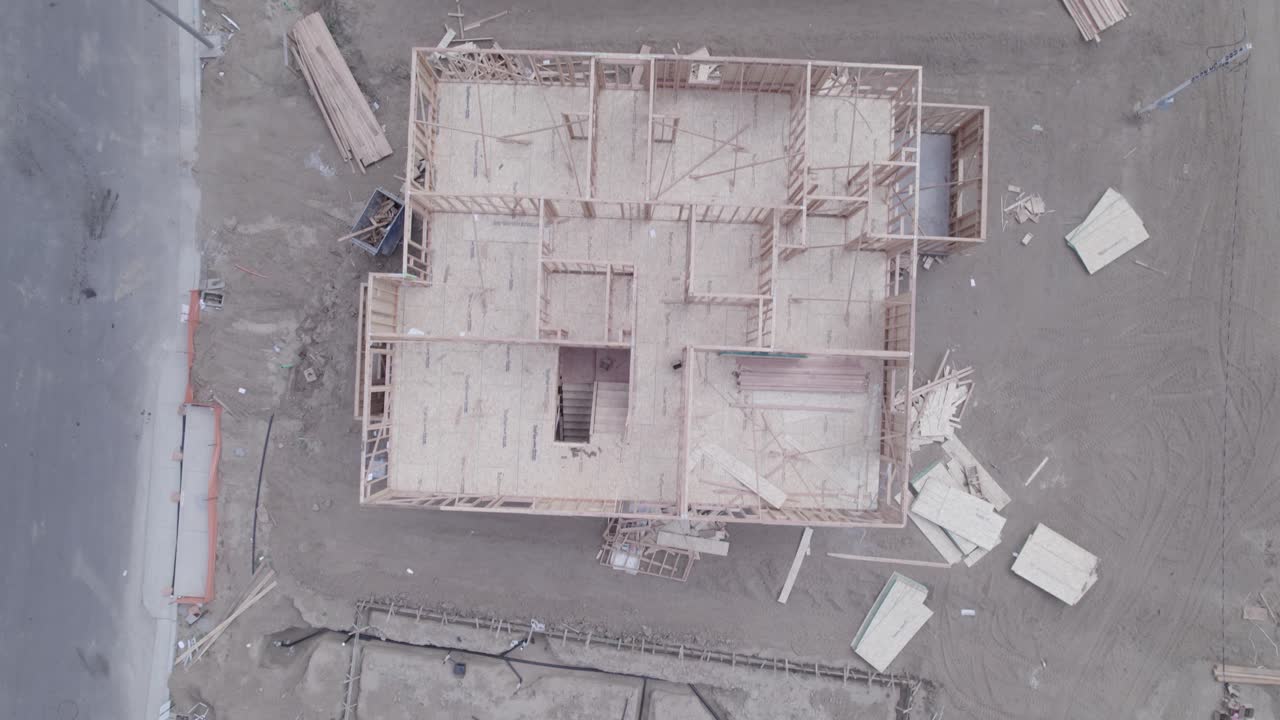 un dron mira directamente hacia abajo en varios lotes dentro de un sitio de construcción, revelando cada etapa desde la tierra desnuda sin cimientos hasta una casa parcialmente construida con un techo dispuesto