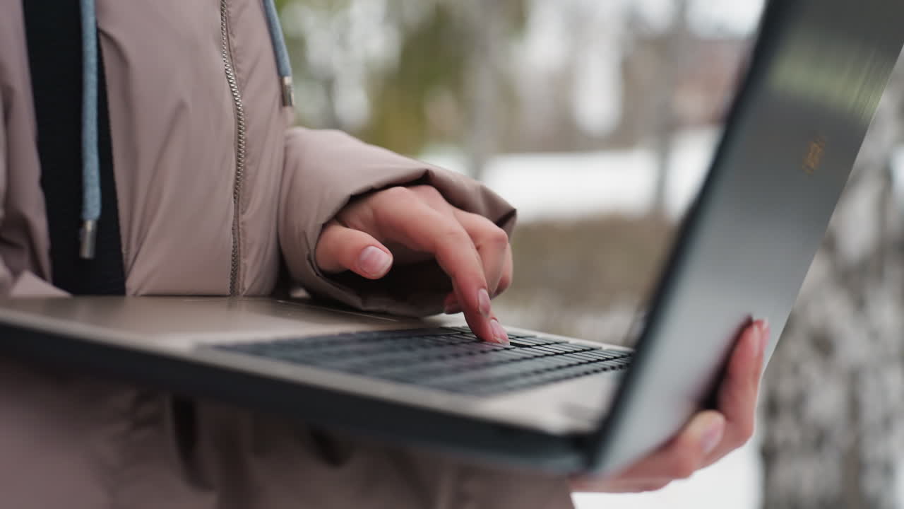 Close-up of light-skinned female hand operating black laptop while holding it with other hand, captured outdoors in winter setting with soft blurred lights in background, showing digital interaction