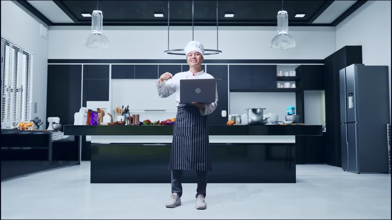 cuerpo lleno de cocinero hombre asiático sonriendo y señalando a una computadora portátil en su mano mientras estaba de pie en la cocina de casa