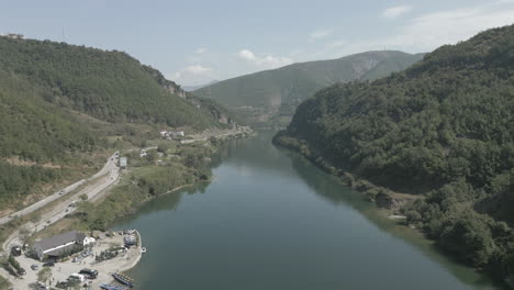 Drohnenschuss-Beim-Flug-über-Die-Fähranlegestelle-In-Fierze,-Albanien,-Mit-Einem-Vorbeifahrenden-Boot-Und-Einer-Fähranlegestelle-Im-Wasser-An-Einem-Sonnigen,-Bewölkten-Tagesprotokoll