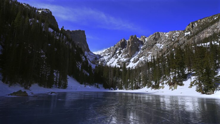 Il lago Dream Lake, ghiacciato d'inverno, nel Parco Nazionale delle Montagne Rocciose (RMNP) in Colorado, sotto un cielo blu soleggiato. Paesaggio mattutino alpino d'alta quota con Hallett Peak, le montagne Flattop, il ghiacciaio Tyndall e i sentieri per Emerald Lake e Bear Lake.