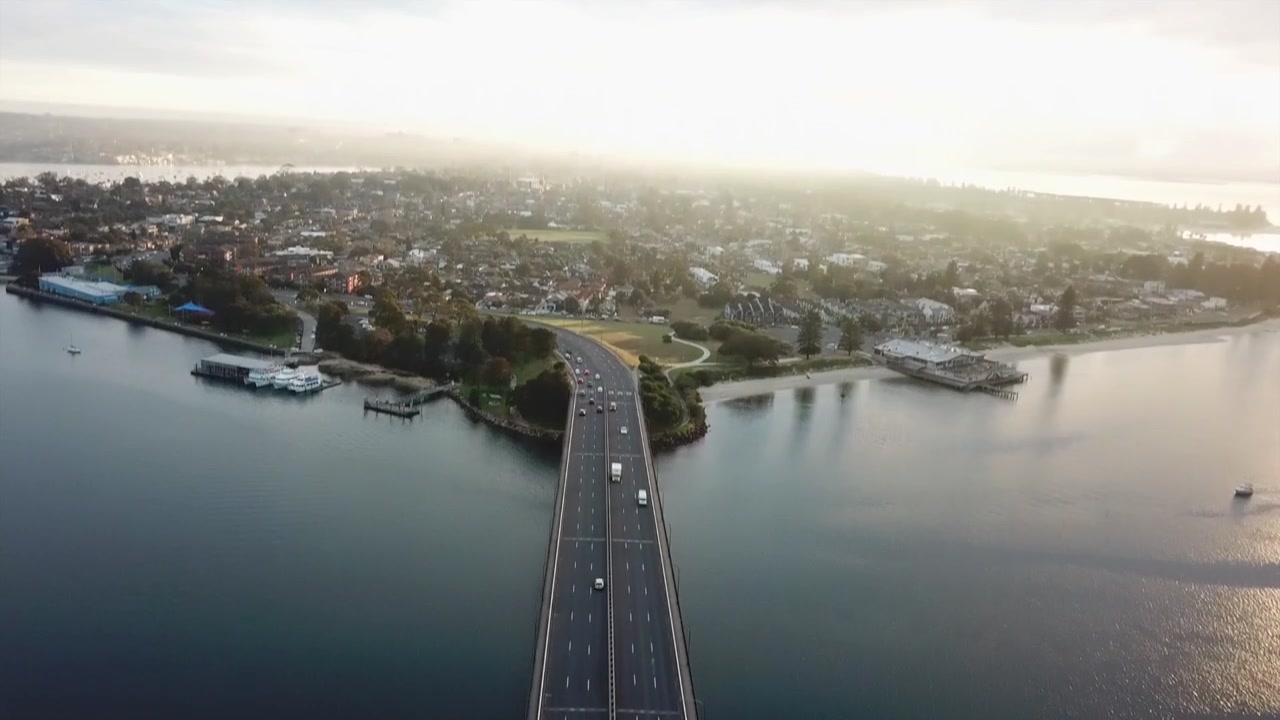 Aerea: Ripresa drone a volo d'uccello su un ponte segue un'auto che si dirige verso la città mentre il sole sorge a Sydney, Australia