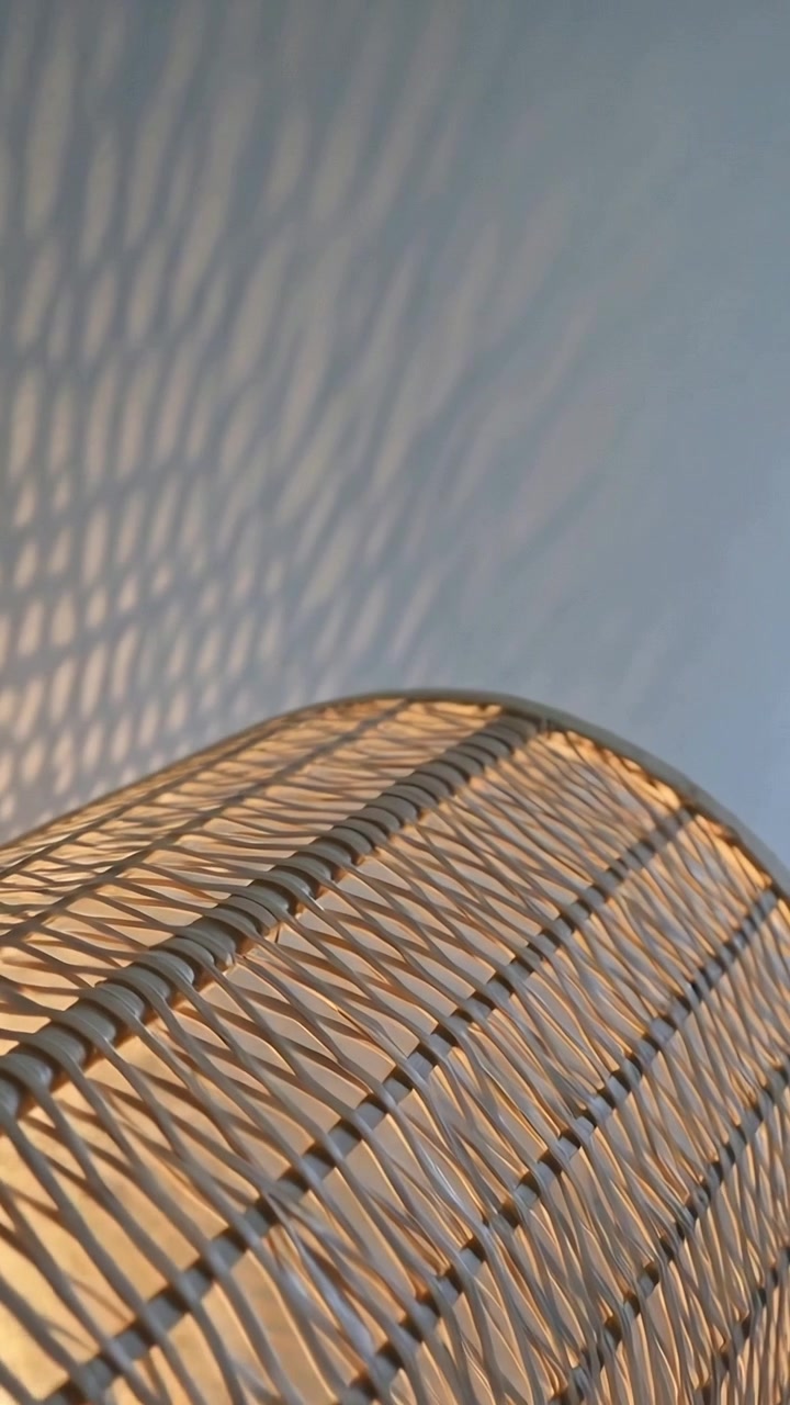 Video verticale: Telecamera inclinata che mostra una lampada in rattan proiettare ombre intrecciate su una parete chiara, accanto a gradini in legno.