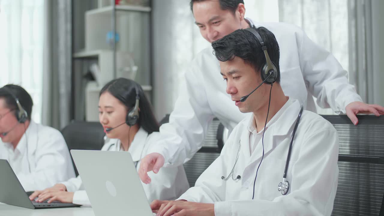 A Man Coming To Help One Man Of Three Asian Doctors In Uniform Using Computer For Working As Call Center Agent While His Colleagues Are Speaking And Typing During  A At The Office
