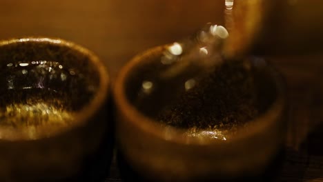 close-up of liquid being poured into two brown ceramic cups on a wooden surface.