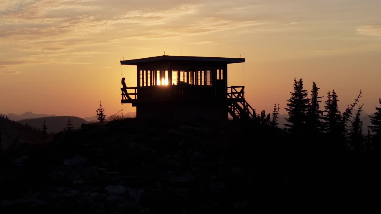 Ripresa aerea con drone in panning che si muove da destra a sinistra, mostrando una torre di avvistamento incendi remota e isolata in silhouette all'alba in Idaho, mentre il sole filtra attraverso le finestre e una donna si appoggia a una ringhiera